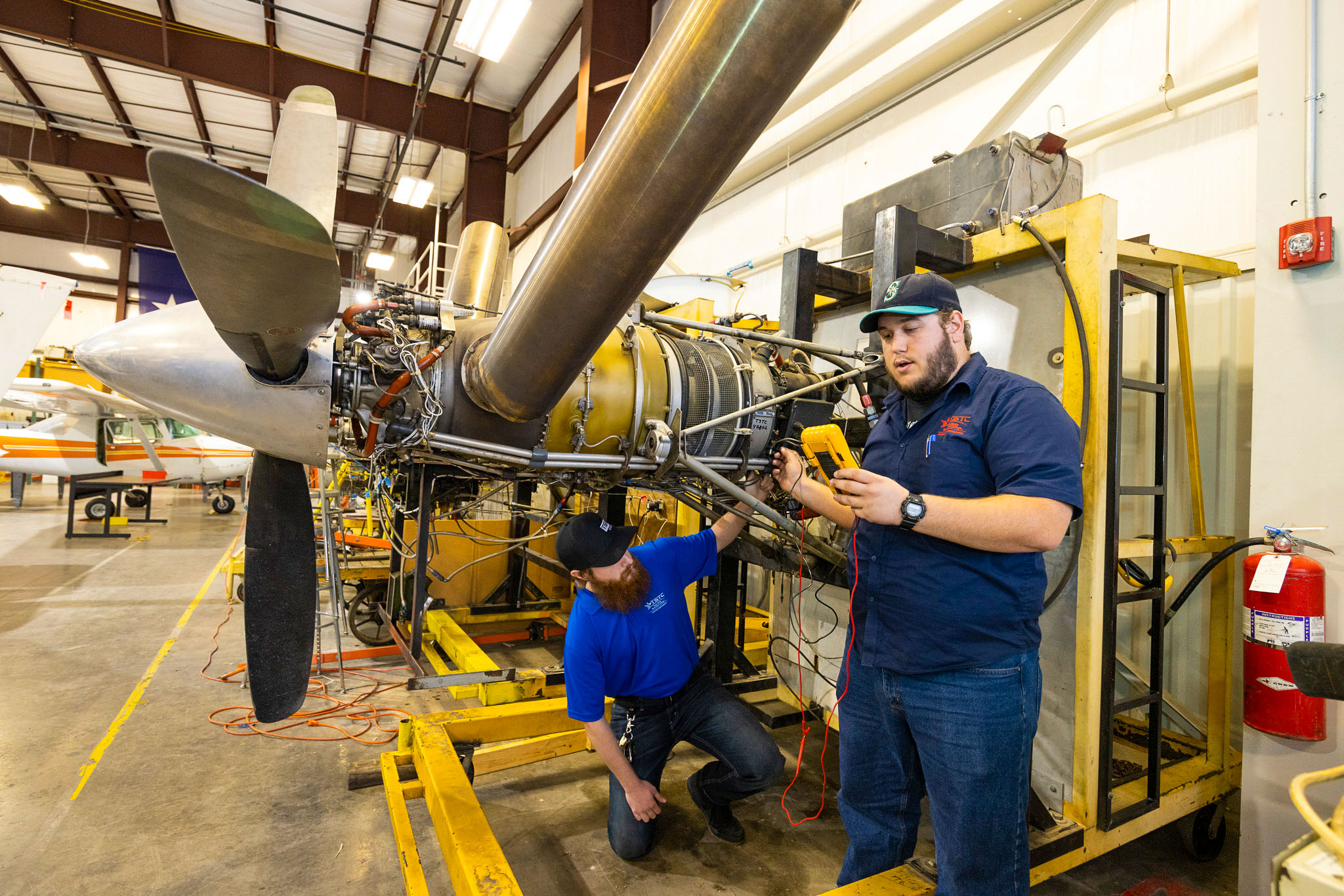 Two aviation students inspect a mounted aircraft engine; one kneels beneath the frame while the other uses a handheld multimeter to test connections beside a large propeller in a hangar-like workshop.